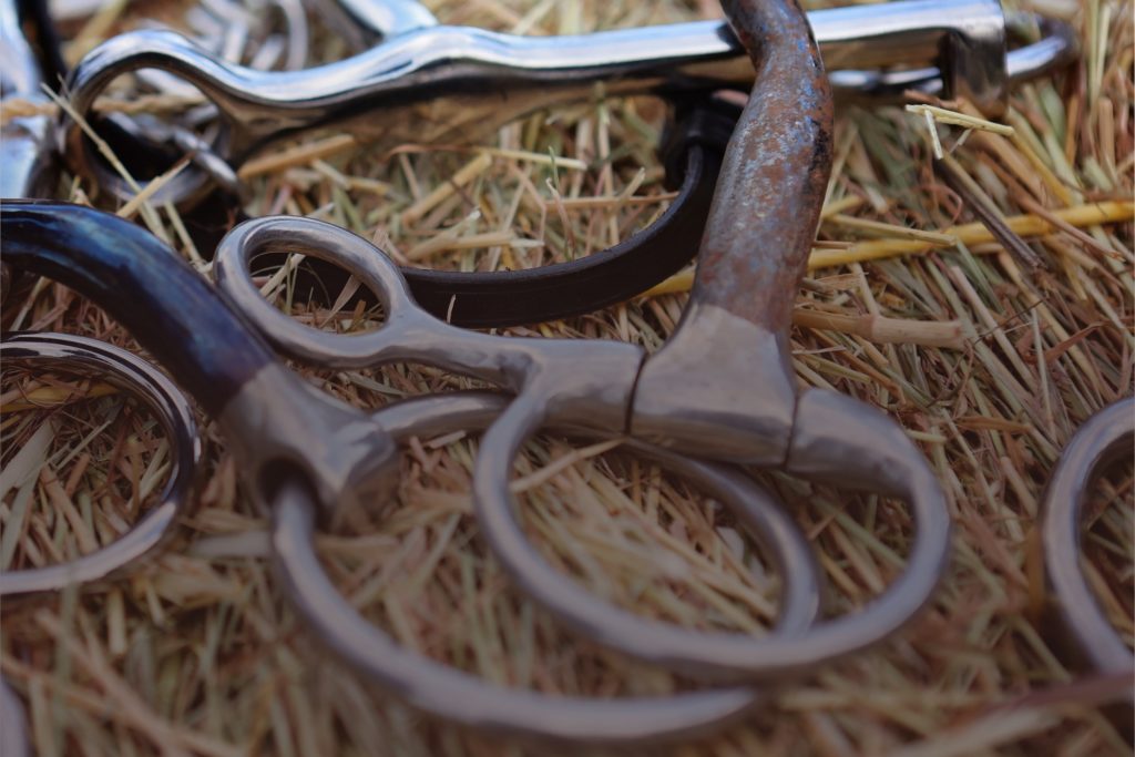 A jumble of bits with different types of mouthpiece and cheeks laying jumbled on a bale of hay